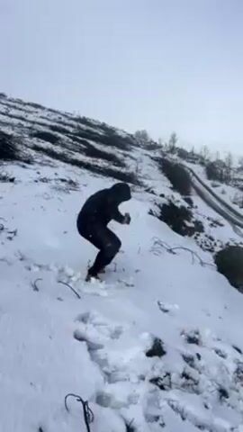 A person dressed in black makes a snowball and throws it in a snowy landscape. The person squats to pack the snowball together and then stands to throw it. A light blue car is visible behind the person on a snowy road, and the surrounding terrain is covered in snow with sparse vegetation. A rainbow is visible in the background.