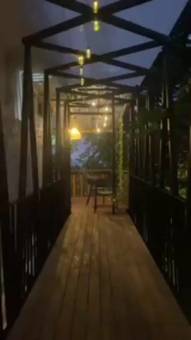 The video presents a view of a covered wooden walkway leading to an outdoor deck area. The walkway is constructed with a black metal frame forming a series of connected geometric shapes overhead, covered with a transparent material. String lights are hung within the frame, providing illumination. The sides of the walkway are bordered by black railings. The wooden planks of the walkway are slightly wet and reflect the ambient light. At the end of the walkway is a deck area with a wooden railing. A chair is positioned on the deck, facing outward, towards a view of trees and the twilight sky. The overall atmosphere is calm and serene, with soft lighting adding to the ambiance. Vegetation is visible to the sides, contributing to the outdoor setting.