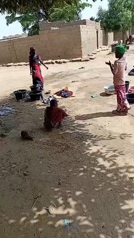 The video shows a scene in what appears to be a village setting. Three individuals, likely women or girls, are engaged in washing clothes outdoors. They are working near several black buckets and a white basin, with clothes scattered on the ground. One person in a green head covering is standing and appears to be directing or observing the others. Two other people, wearing blue head coverings, are actively washing clothes in the buckets. In the background, there are simple buildings, a tree, and a donkey cart. The ground is mostly dirt and sand. The overall impression is one of a daily chore being performed in a rural environment.