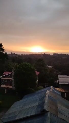 The video shows a sunset over a landscape. The sun is just above the horizon, and the sky is a gradient of orange and yellow. There are trees and buildings in the foreground.  The camera zooms slightly forward.  A child is heard speaking.