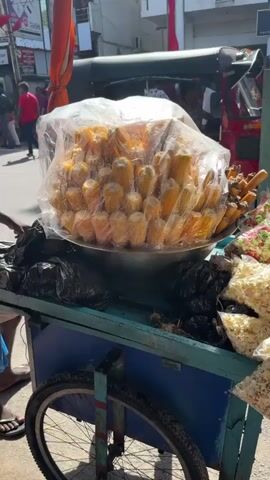 The video captures a vibrant street food scene in Delkanda, Sri Lanka, on a sunny day. The primary focus is a blue street food cart, equipped with two wheels, offering various snacks to passersby.

Dominating the cart is a large metal bowl filled with numerous ears of corn on the cob, each individually wrapped in clear plastic and stacked high, creating a mound. Beside the corn, several small, clear plastic bags filled with popcorn are displayed. Some bags contain plain white popcorn, while others hold colorful popcorn, featuring pink and green kernels. Black plastic bags, possibly for waste or other vendor supplies, are also visible on the cart.

In the background, a bustling street is active with people walking, some wearing red attire, and a red auto-rickshaw (tuk-tuk) is parked. Red flags or banners are visible hanging on buildings along the street, suggesting a possible public event or rally. Buildings line the street, and a sign for "Alaina Food Center" is visible in the distance, along with a partially visible "FACTORY OUTLET" sign. The scene depicts a typical busy day in a South Asian urban environment, with the street vendor playing an integral role in the local commerce and daily life.