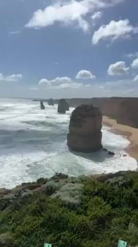 The video shows a scenic view of the Twelve Apostles rock formations in Australia. The stacks stand tall in the ocean, with waves crashing around them. A sandy beach is visible in the background. The sky is blue with scattered white clouds. Green vegetation covers the foreground. The wind is strong, creating significant noise.