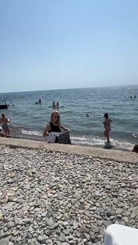 The video shows a busy beach scene on a sunny day. The foreground is dominated by a wide expanse of gray and white pebbles. A concrete walkway runs along the edge of the pebble beach, separating it from the sea. In the middle ground, a few people are visible near the water. On the left, a woman with blonde hair wearing a black swimsuit sits on the concrete, holding a white towel. Further out in the water, other beachgoers are swimming, wading, and some are on paddleboards. The water is blue and calm, with small waves gently lapping the shore. Towards the right side of the frame, a wooden pier extends into the sea, with several people standing on it and some sitting on chairs. The sky is clear and bright blue, indicating fair weather.