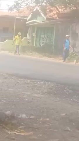 A yellow truck drives down a dusty road, kicking up a cloud of dust. A man wearing a yellow shirt and a hat walks on the side of the road, away from the truck. Two other men, one in a blue shirt and the other in a gray shirt, walk further down the road in the same direction. A building with a red tiled roof is visible behind the men. There is also a green signpost with text on it. The road appears to be paved, and there is vegetation on the side. The overall scene is hazy due to the dust.
