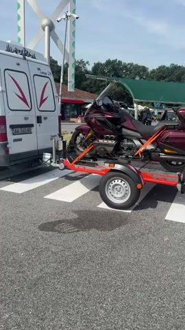 The video shows a white van with red logos, which appears to be a cargo van, parked on a pedestrian crossing with white stripes. A red motorcycle trailer is attached to the rear of the van. A large, dark red motorcycle is secured to the trailer with orange straps. The trailer has a license plate with the characters XA 202MH. In the background, there is a building with a sign that says "AUTOGRILL." There are trees and a clear blue sky. A tall, slender pole with a white, fan-like structure at the top is also visible. The video is a short, static shot.