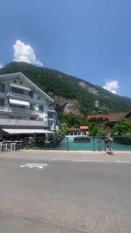 The video captures a sunny day in Unterseen, Switzerland, with a focus on the turquoise Aare River. The scene opens with a view of the river and a building with balconies, backed by a lush green mountain. A person walks across a bridge, and the camera pans to show more of the river and surrounding architecture, including a light-colored building with a brown roof.

As the video progresses, the camera shifts to a wider shot of the river, showcasing the bright blue water flowing between green banks lined with trees and buildings. Mountains dominate the background under a clear blue sky with scattered white clouds. Another building, possibly a hotel, is visible on the riverbank, with umbrellas set up outside.

The footage then transitions to a close-up of a young woman on the bridge, wearing a blue top and a purple headscarf. She smiles at the camera, suggesting a relaxed and happy mood. The camera then pans back to a view of the river and the bridge railing, which has white graffiti on it. The overall impression is a scenic and pleasant travelogue, highlighting the natural beauty and charming atmosphere of the location. The time of day appears to be midday, given the bright sunlight and clear skies.