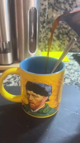 A close-up shot shows coffee being poured from a dark metal pot into a yellow mug. The mug features a painting of a man's face, possibly Vincent van Gogh's self-portrait. A silver spoon rests inside the mug, and a pepper grinder is visible in the background to the left. The coffee stream is dark brown and fills the mug.