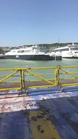 The video shows a harbor with two ferries in the water. The ferry on the left is labeled "DFDS". The ferry on the right is white with green accents. In the foreground, there is a yellow railing with a blue and weathered deck. In the background, there is a white cliff face with a structure on top. The sky is clear and blue. The video pans slightly to the right.