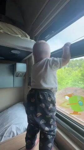 A baby is standing and looking out of a window on a train. The baby is wearing a white t-shirt and dark pants with a pattern of cartoon animals. The train window has stickers of a cartoon astronaut and a rocket ship. Outside the window, there are trees and a train track. There is a bunk bed visible in the background, with a pillow on the lower bunk.