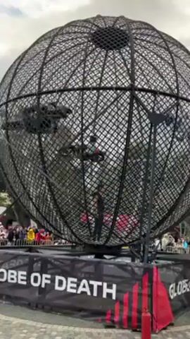 A large black steel mesh sphere labeled "GLOBE OF DEATH" encloses a motorcycle stunt show. Two riders are within the sphere. One is wearing a white and red outfit with a blue helmet and the other is wearing all black. They ride in circles, traveling up the inner surface of the sphere. A crowd watches from behind a barrier. A red fire extinguisher stands on the ground in front of the sphere. The sky is overcast.