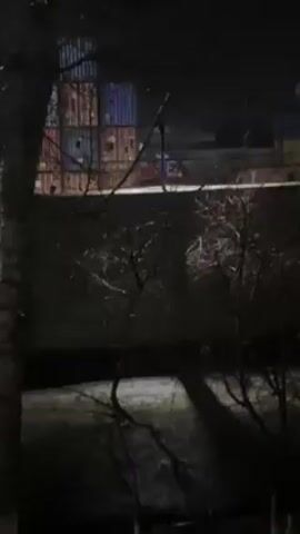The video shows a dimly lit, industrial scene at night. In the foreground, branches of a bare tree are visible. Behind a low brick wall are stacks of cargo containers, some painted in blue, red, yellow, and green. A red vehicle, possibly a truck, can be seen partially behind the wall. It is raining or drizzling as the ground appears wet and reflective. The scene is dark, illuminated by artificial lights that cast indistinct shadows.