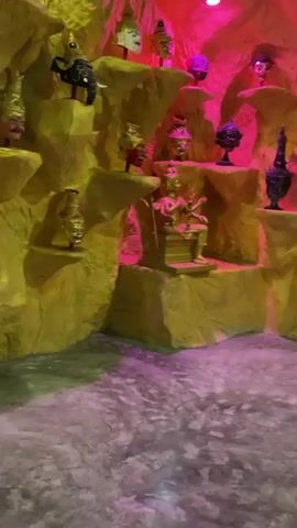 The video pans across a display of traditional Asian puppets or masks, set against a backdrop resembling a cave painted in shades of yellow and pink. Many of the masks depict characters with exaggerated facial features, ornate headdresses, and vibrant colors. Some masks are mounted on pedestals, while others are incorporated into the cave-like structure. In one segment, a puppet appears to be holding a cigarette. The lighting is dramatic, with pink and yellow hues creating an otherworldly atmosphere. The ground appears to be covered in a rough, textured material, possibly representing stone or earth.