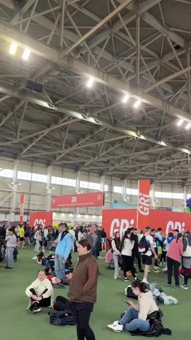 The video shows a large indoor event, possibly a marathon or sports gathering, with numerous people milling around. The event is taking place in a large, well-lit space with a high ceiling featuring exposed metal beams. Many participants are wearing athletic clothing. Some are standing, others are sitting or lying on what appears to be a green artificial turf. Backdrops with "GRI" and other words are visible, possibly representing the event's name or sponsor. There are also balloon decorations, which are green, purple, and red. People are mostly walking, standing, resting, or using their phones.