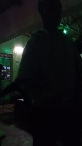 The video shows two people dancing in a dimly lit room, possibly a bar or club. The lighting is provided by colorful lights and a ceiling fixture. The walls of the room appear to be painted green.  The dancers are silhouetted, but one person wears a white t-shirt.  Tables and chairs are visible in the background.