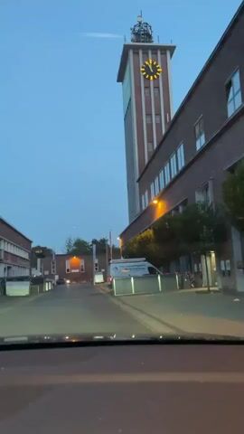 The video shows a shot from a vehicle driving down a road towards a tall, brick building with a clock tower. The sky is a deep blue, indicating either early morning or evening. The clock on the tower displays approximately 5:10. Other brick buildings line the road, and there are streetlights and trees. A white van with lettering is parked on the right side of the road, and a few cars are visible in the distance. The overall impression is of an industrial or institutional setting.