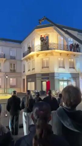 Here's a description of the video:

The video shows a stunt involving two people performing on a set resembling a European street scene at night. The backdrop includes buildings with balconies.

Two individuals, wearing dark clothing and red helmets, are connected to wires and are performing a stunt. One person is thrown off a balcony while the other pulls the first one up using the wires, creating an aerial effect. 

A group of people are standing below, watching and recording the stunt. A crane with a camera is visible in the background, suggesting the scene is being filmed professionally. The sky is dark blue, indicating it is dusk or night. The overall impression is that this is a scene from a movie or theatrical performance.