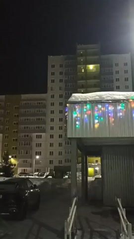 The video presents an exterior nighttime view of a residential area, possibly a housing complex in a cold climate, as suggested by the presence of snow. A multi-story apartment building with numerous illuminated windows dominates the background, rising vertically from the lower left to the upper right of the frame. To the right of the building, a structure with a metallic exterior is decorated with colorful string lights, lending a festive air to the otherwise stark setting. The roof of this structure is covered in a layer of snow.

A black car is parked in the foreground to the left, partially obscured by the shadow cast by the structure. Bare trees and a few smaller buildings can be spotted further back. Streetlights cast a bright glow, adding to the artificial illumination of the scene. The sky above is dark and clear, suggesting a cold night. The overall composition captures a quiet urban environment during the winter season.