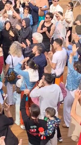 A crowd of people, including men, women, and children, is gathered on what appears to be a ship's deck. They are clapping, cheering, and raising their hands in the air. People are wearing a variety of casual clothing, including T-shirts, tank tops, jackets, and jeans. There are a few people wearing sunglasses. The deck has wooden planking. Other people are walking past in the background, seemingly unaffected by the celebration. The environment suggests they may be on a cruise or ferry.
