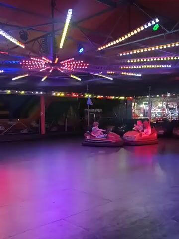 The video shows a bumper car ride at night. There are several bumper cars on the floor, two of which are visible with people inside. The cars are red and beige, and they are illuminated with colorful lights. The floor is dark and reflective, and it is lit with purple and pink lights. The ceiling is decorated with many colorful lights arranged in a radial pattern, and there are also some strings of lights in rows. In the background, there are some booths or attractions that are also lit up with lights. The people in the bumper cars appear to be enjoying themselves, and the overall atmosphere is festive and energetic.
