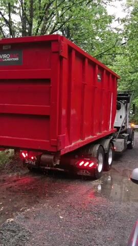 This media file captures a bright red dumpster truck, its bed tilted slightly upwards, in Saint-Colomban, Canada. The truck is positioned on a gravelly, wet road, suggesting recent rain or damp conditions. The surrounding environment is lush and green, with trees visible in the background, indicating an outdoor, possibly rural or suburban setting.

The dumpster itself is predominantly red and features a logo with the text "ENVIROBAC LAURENTIDES". The truck's rear lights are illuminated, showing red brake lights and amber turn signals, though no immediate action is apparent. The license plate is visible and reads "LB52011". The overall impression is a functional vehicle, likely engaged in waste removal or a similar service, operating in a natural, somewhat overcast setting. There is no clear indication of the time of day, but the lighting suggests it's daytime. The scene is static, with no people clearly visible in the frame. The mood is neutral, focusing on the industrial equipment within its natural environment.