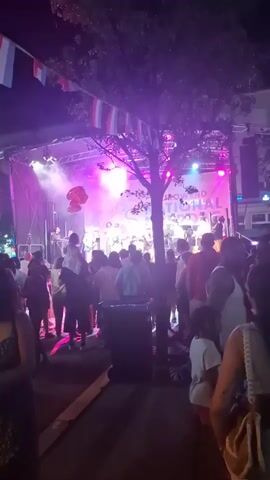 This is a video taken at an outdoor event at night. A band is performing on a stage under bright pink and white lights. A banner behind the band reads "POWER TO THE PEOPLE". A large, balloon-like object resembling a mushroom is floating to the left of the stage. A crowd of people are standing in front of the stage, watching the performance. Some people are dancing, while others are standing and talking. There are trees with leaves in the foreground. Strings of triangular flags are draped across the top of the scene. The overall atmosphere appears to be festive.