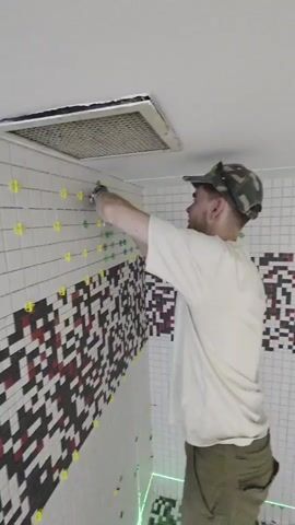 A man wearing a camouflage baseball cap and a light-colored t-shirt is tiling a wall. He is holding a tool to place tiles. The wall is partially tiled with white tiles and has a pixelated pattern of black, white, and red tiles. Yellow spacers are visible between some of the tiles. A ventilation grate is installed in the ceiling above the tiled wall. The room appears to be under construction, with a green laser line visible on the floor and buckets in the background.