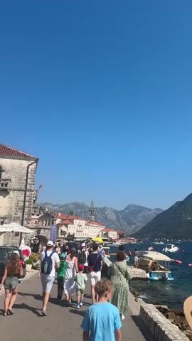 A group of people are walking along a paved walkway next to a body of water. On the left side of the walkway, there is a stone building with a red-tiled roof and a flag. Further down the walkway, there are shops and restaurants with umbrellas and awnings. In the background, there are mountains and a church tower. On the right side of the walkway, there are boats docked in the water. The sky is clear and blue.