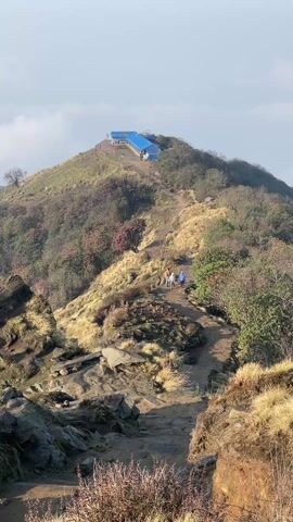 The video shows a group of four people hiking uphill on a narrow trail on a mountain. The trail is rocky and dirt-covered, with grass and shrubs on either side. In the distance, at the top of the mountain, is a building with a bright blue roof. The sky is overcast. Birds are chirping in the background.