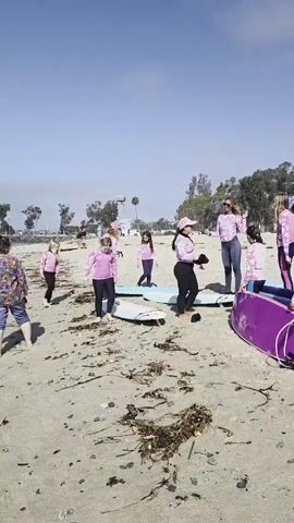 The video shows a group of young girls on a sandy beach with surfboards. They are dressed in matching pink long-sleeved rash guards and some are wearing pink hats. The surfboards are of various colors, including purple, turquoise, and white. Some of the girls are standing, while others are sitting or crouching near the surfboards. The environment is sunny with a blue sky and scattered clouds. Trees and some structures are visible in the background. The girls appear to be preparing for or having just finished a surfing lesson or activity.
