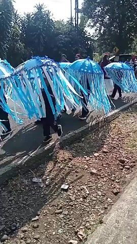 The video shows a parade or procession taking place outdoors on a paved road. Several people, dressed in black clothing, are walking and carrying large, umbrella-like structures that are decorated to resemble jellyfish. These jellyfish costumes are blue and white, with long, flowing streamers that hang down. The people are walking in a line, and the costumes create a colorful and dynamic visual effect. In the background, trees and foliage are visible, suggesting a rural or suburban setting. A motorcycle passes by in the middle ground. The ground alongside the road is covered in gravel and dirt.