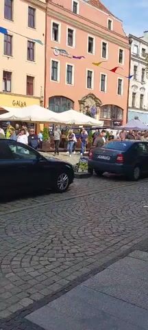The video shows a cobblestone street scene with buildings lining one side. Several people are visible, some standing and talking, others walking. Two cars are parked on the street. White umbrellas suggest outdoor seating or market stalls. Flags or banners are strung across the buildings. The overall atmosphere appears to be a daytime gathering or market event.