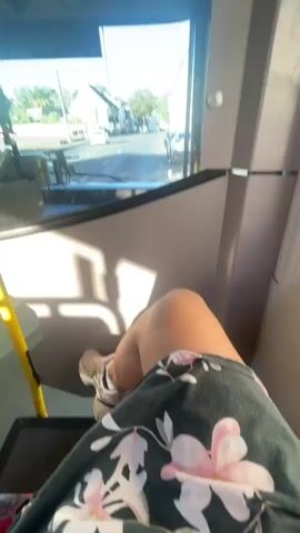 A person's legs are visible in the foreground, wearing a floral-patterned skirt and white sneakers. They are seated on a bus, with their legs extended. The view from the bus window shows a street with cars and trees. A cyclist is visible riding down the street. The sun is shining, casting shadows on the road. The bus appears to be moving.