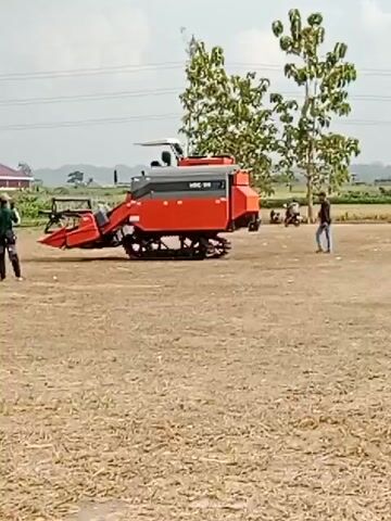 A group of people stand near a large orange and gray combine harvester with tracks. The machine is parked on a dry, flat, dirt or grass area. In the background, there are trees, a building, and a hazy sky. One person in a green shirt and dark pants stands to the left of the combine, facing it. Another person in a blue shirt and dark pants stands to the right of the combine, facing away from it. Two other people are visible in the distance. The combine appears to be in a field. The video is shaky, suggesting it was filmed by someone holding a phone.