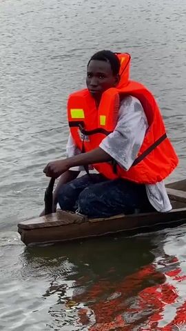A young man wearing an orange life jacket over a white shirt and blue pants is sitting in a wooden canoe and paddling with an oar. He is looking forward. Another person, also wearing an orange life jacket, is standing in the canoe behind him. They are on a body of water with vegetation on the shore in the background. The water is rippling.