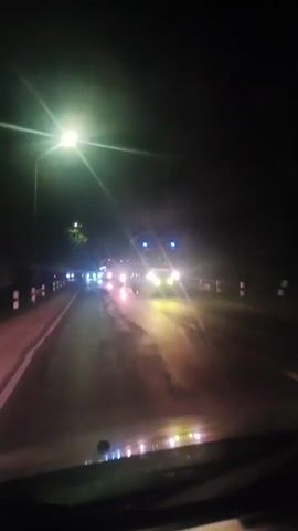 A first-person perspective video shows a nighttime drive on a road. Streetlights and the headlights of oncoming vehicles illuminate the scene. The road appears wet, reflecting the lights. Several vehicles, including cars and what appears to be a truck or bus, are visible in the distance. The video captures the movement of the car as it travels along the road, with the landscape blurring into streaks of light due to the motion and low light conditions.