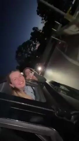 The video shows two young women in a car at night. One woman is partially out of the sunroof, looking up and laughing while covering her mouth with her hand. The other woman is inside the car, holding a phone, possibly recording. The car appears to be at a gas station or similar roadside stop, as there are signs and lights visible in the background. The scene is dimly lit, with the car's headlights illuminating the immediate surroundings.