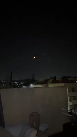 A video shows a dark sky with a glowing orange object floating upwards. The object appears to be a sky lantern. Below the sky, there is a cityscape with buildings and lights. The foreground shows a balcony with a wall and a decorative pot. The video is very short and shows the lantern slowly ascending into the night sky.
