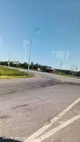 The video shows a view from inside a vehicle, looking out at a rural road intersection on a clear, sunny day. The road surface is asphalt and appears worn in places, with visible white lane markings. A bus stop shelter, painted red and white, is situated at the intersection, along with a lamppost and utility poles with wires. The surrounding landscape consists of a treeline and a vast expanse of clear blue sky. The vehicle's interior is partially visible through reflections on the glass, indicating the perspective of someone traveling. The overall impression is one of a quiet, open, and somewhat isolated location.