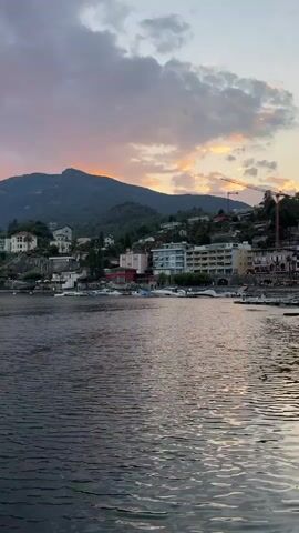 A static video captures a wide shot of a tranquil lake with a town nestled along its hilly shoreline. The sky above displays a gradient of soft pink and orange hues, indicative of a sunset or sunrise, contrasting with the muted gray clouds. Buildings, ranging from white multi-story structures to smaller houses, are visible cascading down the hillside, interspersed with lush green trees. Several boats are docked along the shore, and a lone sailboat glides across the lake in the mid-ground. The water's surface is gently rippled, reflecting the subdued light from the sky. The overall atmosphere is serene and picturesque, with a crane visible near the town, suggesting ongoing development or maintenance. No people or animals are discernible within the frame.