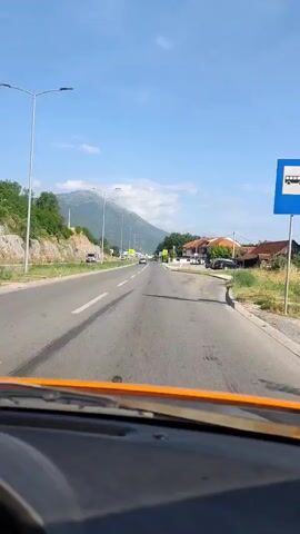 The video captures a forward-facing view from inside a moving vehicle traveling along a paved road on a bright, sunny day. The sky is largely clear blue with a few scattered white clouds.

On the left side of the road, there is a green, rocky embankment, with a white van parked on the shoulder further ahead. Modern streetlights with long arms line both sides of the road. In the distance, a large mountain, partially obscured by light haze or clouds, forms the backdrop.

To the right of the road, a collection of low-rise buildings with distinctive red-tiled roofs and white facades are visible, set among trees and green vegetation. Several cars are parked on the side of the road near these structures.

The road itself is multi-laned, with clear white solid and dashed lane markings, as well as yellow markings on the right-hand shoulder. Other vehicles can be seen driving ahead in the distance. Two prominent yellow triangular warning signs, each featuring a black exclamation mark, are visible on both sides of the road further down.

The lower part of the frame reveals the orange dashboard of the recording vehicle. The overall scene suggests a journey through a semi-urban or rural area, consistent with the given location of Danilovgrad, Montenegro.