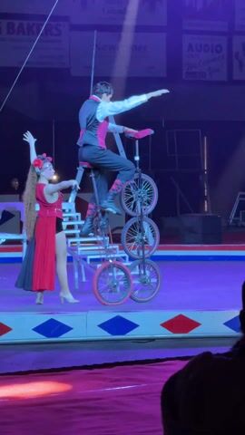 The video captures a circus performance featuring two acrobats on a purple circular stage.

In the foreground, a female performer with long, flowing red hair and a red floral headpiece is seen. She is wearing a sleeveless red dress with black shorts underneath and black boots. Her left arm is raised in a performance pose, while her right hand is initially assisting the male performer.

The male performer is balanced on a tall, multi-wheeled unicycle. He is dressed in a white shirt, a black vest with a pink/red trim, black pants with pink/red accents on the lower legs, and black boots. The unicycle is unusual, appearing to be a stack of three wheels, one directly above the other, making it exceptionally tall. He is positioned on the seat of the topmost wheel.

At the beginning of the video, the female performer is holding onto the unicycle's frame, helping to steady it. As the video progresses, she releases her grip and moves slightly away, leaving the male performer to balance independently. He maintains his balance by extending his arms outwards, demonstrating control and skill on the elevated unicycle.

Behind the performers, there's a white-colored structure that resembles a set of stairs or scaffolding, adding to the stage design. The overall lighting is dramatic, with purple and blue hues illuminating the stage, and a bright spotlight focused on the performers. In the darker background, parts of signs are visible, including "BAKERS," "Newcomer," and "AUDIO." The stage itself has a distinctive red, white, and blue checkered border. The audience's heads are visible in the very bottom right corner of the frame, indicating an indoor performance setting.