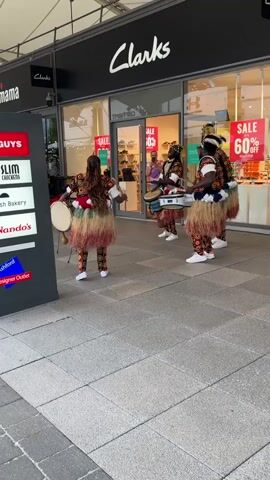 Four people play drums in front of a Clarks shoe store. They wear matching patterned outfits with a grass skirt and hats with feathers. The man on the left plays a tall round drum while the others play smaller drums that hang from their necks. Signs in the background include "mama," "GUYS," "SLIM CHICKENS," "FRESH Bakery," "Nando's" and "Ashford Designer Outlet." The Clarks store has signs advertising a sale of up to 60% off. Customers are visible in the store. The ground is paved with gray rectangular tiles.