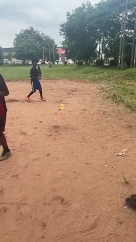 A group of young men are engaged in a training session on a dirt field. One man in a black t-shirt and blue shorts is performing agility drills, running around cones placed on the ground. Another man in a yellow shirt with black patterns is in the background, also seemingly participating in the training. In the foreground, a man in a dark blue t-shirt and dark shorts is about to kick a ball towards the man in the black t-shirt. The environment appears to be an outdoor sports ground with trees and buildings in the background, under an overcast sky.