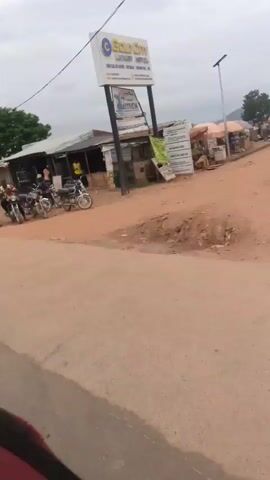 The video shows an outdoor scene with a dirt road in the foreground. Several motorcycles are parked near a row of buildings that appear to be shops or small businesses. Some people are standing or sitting around the motorcycles and buildings. One person is riding a motorcycle and moving from left to right across the frame. The sky is overcast. A sign reading "CAR WASH" is visible on one of the buildings. Another sign with text that is difficult to read is also present. The overall impression is of a casual gathering or stopping point in a semi-urban or rural environment.