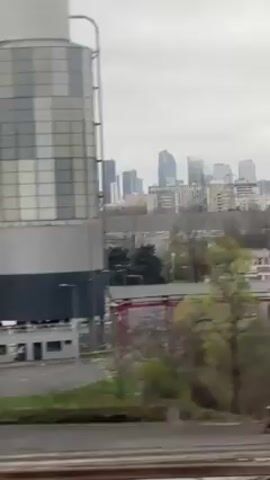 The video shows a view from a moving train. In the foreground are two large, cylindrical structures with a silver and gray checkered pattern. Below them is a fenced area with several vehicles, including tanker trucks and vans. In the background, a city skyline is visible, featuring several tall buildings. The sky is overcast and gray. There is a strip of green grass along the bottom of the frame, and railroad tracks are visible. The overall setting appears to be an industrial area on the outskirts of a city.