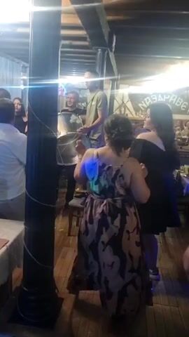 The scene depicts a lively indoor celebration or party. Multiple individuals are present, some dancing and others engaging in conversation. At least two women are dancing, holding hands. A man can be seen playing a drum, with another person assisting him in positioning it. A laptop and other equipment indicate a DJ setup. The room has a rustic feel, with wooden beams and a sign on the wall in a foreign language. The lighting is bright, with additional colored light effects adding to the celebratory atmosphere.