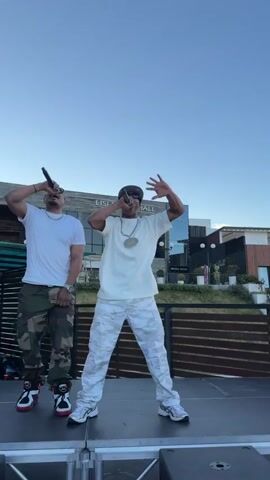 This is a video of a musical performance. Two men are on a raised stage, holding microphones and performing. The man on the left is wearing a white t-shirt and camouflage pants. The man on the right is wearing a white t-shirt, white camouflage pants, sunglasses, and a large gold chain. Behind them, a building with "LISI EVENT HALL" visible on it. Another person is visible in the background holding up a phone, seemingly recording the performance. The sky is clear and blue. The stage appears to be outdoors, with some wooden fencing and greenery visible.