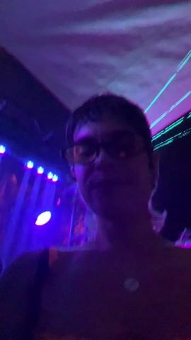 Two women are in a dark club, lit by blue spotlights and green laser beams. The woman on the right smiles at the camera and wears sunglasses, a black and orange top, and a pendant necklace. The woman on the left has long hair and is wearing a patterned top and a necklace. They are surrounded by people and the sound of electronic dance music.