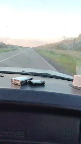 The video shows the dashboard of a car driving down a road. A pack of cigarettes and a lighter are visible on the dashboard. The road is paved and has a grassy shoulder. Trees and vegetation are visible on either side of the road. The sky is clear and the sun appears to be setting. The car's dashboard has the "Audi 80" logo.