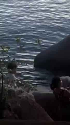 Two individuals are positioned on a rocky shoreline alongside a body of water. The person on the left is partially obscured by foliage, while the person on the right is visible, seated on a rock. A large boulder lies near the water's edge. The water itself shows gentle ripples.